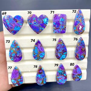 Könnte beinhalten: Eine Sammlung polierter Edelsteine in verschiedenen Formen, darunter Herzen und Tropfen. Die Steine weisen eine lebendige Mischung aus lila, blau und goldfarben auf. Jeder Stein ist zur Identifizierung nummeriert und wird auf einer weißen Oberfläche präsentiert.