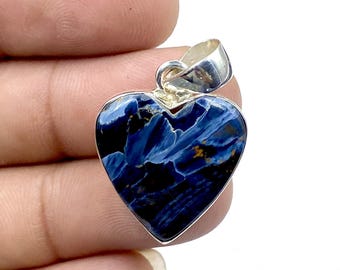 May include: A heart-shaped pendant featuring a polished blue and black stone with subtle golden-brown inclusions, set in a smooth silver frame with a silver bail at the top for attachment to a chain or cord, showcasing a smooth, reflective surface.