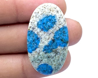 May include: Oval-shaped K2 stone, featuring a speckled gray and white base with vibrant blue patches. The polished gemstone displays a natural, textured appearance, showcasing a unique pattern and color contrast.