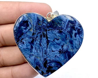 May include: A heart-shaped pendant featuring a polished blue pietersite gemstone with swirling dark blue and black patterns. The stone is set in a smooth silver-toned metal frame with a small metal bail attached at the top for hanging.