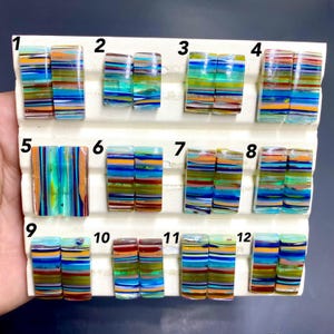 May include: A collection of twelve rectangular glass beads with colorful horizontal stripes in shades of blue, orange, green, and brown. The beads are arranged on a white surface, showcasing their vibrant patterns and textures. Each bead is unique.