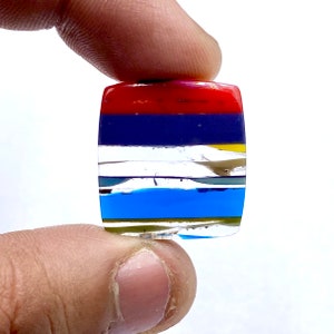 May include: A multi-colored layered stone with red, blue, yellow, and silver stripes. The stone is rectangular and has a smooth, polished surface.
