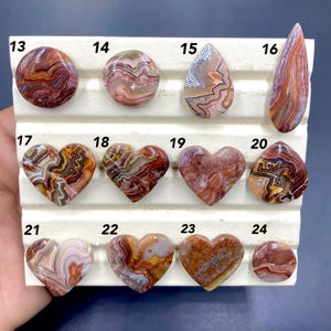 May include: A selection of polished gemstones in diverse shapes, including round, teardrop, and heart. The stones showcase a variety of colours, such as red, brown, white, and beige, with unique patterns and textures. The gemstones are displayed on a white surface.