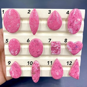 May include: A collection of twelve polished gemstones in various shapes, including oval, teardrop, and heart. The stones are a vibrant pink color with white marbling, set against a white background. Each stone is numbered for identification.