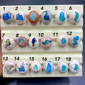 Puede incluir: Una exhibición de dieciocho esferas de ópalo pulidas, cada una con patrones y colores únicos. Los ópalos están colocados sobre un expositor de color crema. Los ópalos presentan una variedad de colores, incluyendo azul, verde, rosa y blanco. Los números 1-18 están impresos encima de cada ópalo.