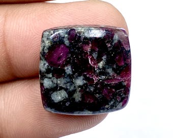 Puede incluir: Una gema de corte cuadrado con una mezcla de colores rojo oscuro y gris. La piedra tiene un aspecto moteado con inclusiones de color rojo intenso. Los bordes son redondeados y la superficie está pulida, lo que le da una textura suave. La gema se sostiene entre los dedos.