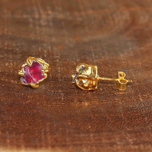 May include: A pair of stud earrings featuring raw, irregular-shaped red gemstones set in textured gold-toned metal. One earring is shown from the front, displaying the deep red stone, while the other is shown from the side, revealing the post backing.