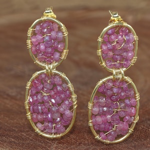 May include: Gold-toned earrings with a double oval design. Each oval is filled with small, irregular pink gemstones, secured by thin gold wire. The earrings have a simple, elegant style.