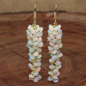 Peut inclure: Une paire de longues boucles d'oreilles pendantes dorées avec des perles d'opale blanche. Les boucles d'oreilles sont composées d'un groupe de petites perles d'opale rondes qui pendent d'un crochet doré.