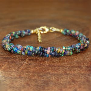 Puede incluir: Una pulsera de ópalo multicolor con un cierre dorado. La pulsera está hecha de pequeñas cuentas redondas de ópalo en tonos azules, verdes, morados y naranjas. La pulsera se muestra sobre una superficie marrón.
