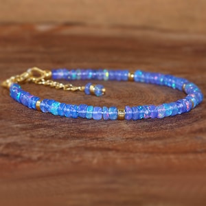 May include: A blue opal beaded bracelet with gold-coloured accents. The bracelet features small, rectangular blue opal beads strung together with gold-coloured metal. The bracelet is displayed on a wooden surface.