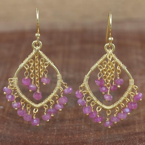 Può includere: Un paio di orecchini color oro con un design geometrico. Gli orecchini presentano una montatura a forma di diamante con catenine pendenti e piccole perline di pietre preziose rosa. Gli orecchini hanno una chiusura a gancio.