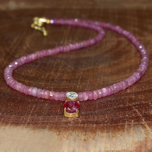 May include: A pink gemstone necklace with a gold pendant featuring a round clear stone and a red gemstone.
