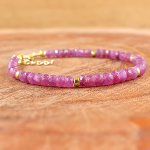 May include: A delicate bracelet made with small, faceted pink gemstones and gold accents. The bracelet is on a wooden surface.