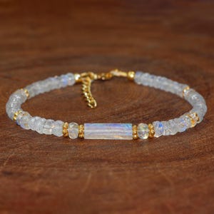 Op de afbeelding: Een delicate armband gemaakt met iriserende witte maansteen kralen en gouden accenten. De armband heeft een grotere rechthoekige maansteen kraal in het midden.