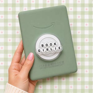 May include: A sage green e-reader with a white pop socket that reads "BOOK GIRLIE" in black letters. The e-reader has the Amazon logo and is held by a person.