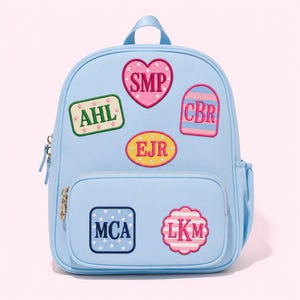 May include: A light blue backpack featuring five embroidered patches with monograms. The patches include a pink heart with SMP, a green rectangle with AHL, a striped arch with CBR, a yellow oval with EJR, a blue square with MCA, and a pink scalloped circle with LKM.