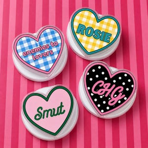 May include: Four heart-shaped coasters with various designs and text. One coaster has a blue and white gingham pattern with the text "enemies to lovers." Another has a yellow and white gingham pattern with the name "Rosie." A third has a black polka dot pattern with the initials "CHG." The last has the word "Smut."