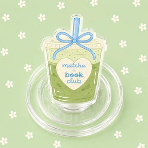 May include: A clear acrylic drink coaster featuring a matcha latte design. The drink is in a clear cup with a blue straw and bow. The words "matcha + book club" are inside a white heart. The coaster is on a clear, round coaster.