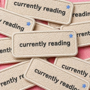 Può includere: Primo piano di toppe in tessuto beige ricamate con le parole "currently reading" in nero. Ogni toppa ha una forma rettangolare con angoli arrotondati e un bordo bianco. Alcune toppe hanno una piccola stella blu. Le toppe sono sparse su una superficie rosa.