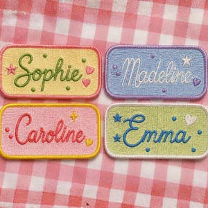 Op de afbeelding: Vier rechthoekige geborduurde patches met afgeronde hoeken. Linksboven is geel met groene tekst Sophie, rechtsboven is blauw met witte tekst Madeline, linksonder is roze met rode tekst Caroline en rechtsonder is lichtgroen met blauwe tekst Emma. Elke patch is voorzien van kleine geborduurde sterretjes, hartjes en stippen.