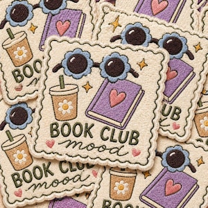 May include: Embroidered patches with a cream-colored background feature a purple book with a pink heart, sunglasses, a coffee drink, and the words "BOOK CLUB mood." The patches have a scalloped edge and are decorated with stars and hearts.
