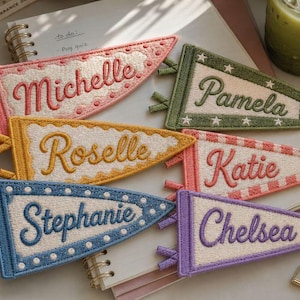 Puede incluir: Seis banderines triangulares de fieltro con los nombres bordados Michelle, Pamela, Roselle, Katie, Stephanie y Chelsea. Cada banderín muestra colores únicos, incluyendo rosa, verde, amarillo, coral, azul y morado con patrones decorativos como estrellas y puntos.