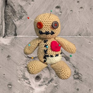 Könnte beinhalten: Handgemachte beige Häkelpuppe mit Knopfaugen und einem roten Herz. Die Puppe hat schwarze Nahtdetails, bunte Nadeln und einen Knopfmund. Die Puppe sitzt auf einer strukturierten grauen Oberfläche.