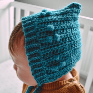 May include: A teal blue crocheted baby bonnet with a textured pattern and pom-poms.