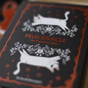 May include: A black box with a white and orange design featuring two white cats and the text "Felis Oracle - The Pawsitive Vibes - "