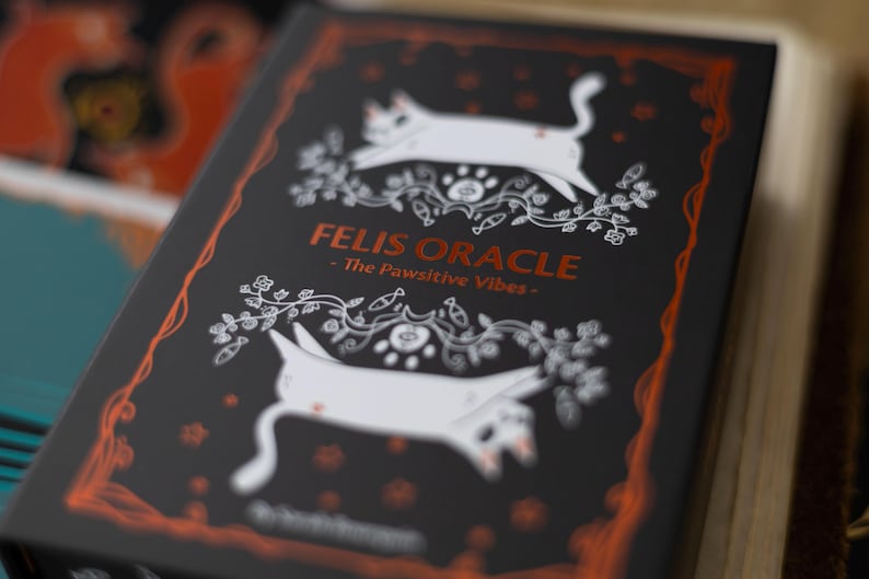 May include: A black box with a white and orange design featuring two white cats and the text "Felis Oracle - The Pawsitive Vibes - "