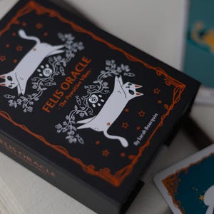 May include: A black box with a white cat illustration and the text "FELIS ORACLE - The Pawsitive Vibes" in orange. The box is surrounded by a decorative orange border.