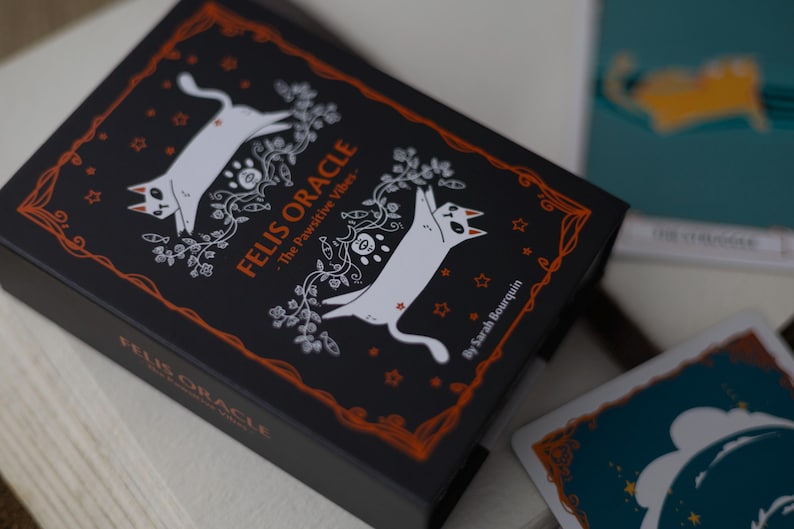 May include: A black box with a white cat illustration and the text "FELIS ORACLE - The Pawsitive Vibes" in orange. The box is surrounded by a decorative orange border.