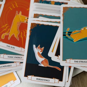 May include: A set of colorful cat-themed tarot cards with illustrations of cats in various poses. The cards feature titles such as "The Sun", "Toxic Energy", "Vulnerable", "Persistence", and "The Struggle".