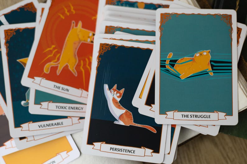 May include: A set of colorful cat-themed tarot cards with illustrations of cats in various poses. The cards feature titles such as "The Sun", "Toxic Energy", "Vulnerable", "Persistence", and "The Struggle".