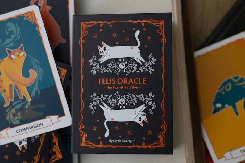 May include: A black card with an orange border features a white cat with a black tail and black ears. The card has the text "Felis Oracle - The Pawsitive Vibes - By Sarah Bourquin" in orange text.