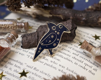 May include: A gold-toned enamel pin featuring a blue bird with gold stars and a crescent moon. The bird is perched on a branch with a book and small bottles in the background.