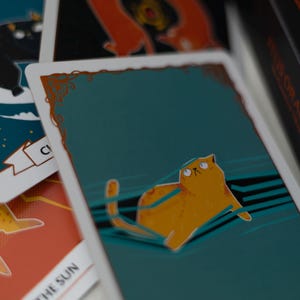 May include: A teal blue tarot card with a gold border and a cartoon orange cat with green eyes looking up. The cat is lying on a blue and white striped surface. The card is part of a tarot deck.