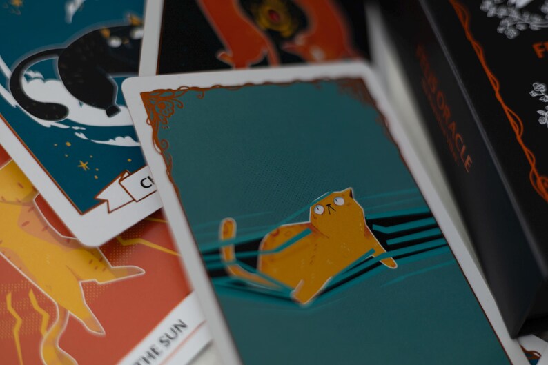 May include: A teal blue tarot card with a gold border and a cartoon orange cat with green eyes looking up. The cat is lying on a blue and white striped surface. The card is part of a tarot deck.
