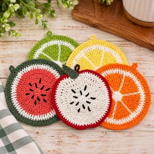 May include: A set of five round, crocheted coasters shaped like fruit slices. The collection includes a green lime, a yellow lemon, a red watermelon, a white apple slice with a green leaf, and an orange slice, each featuring detailed stitching.