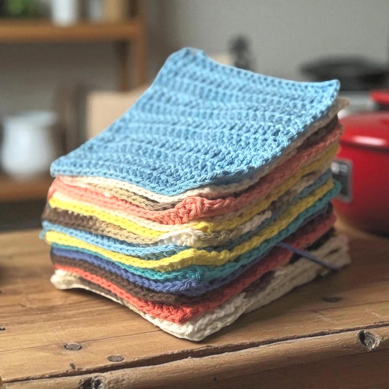 May include: A stack of square crochet washcloths in various colors including light blue, peach, yellow, brown, white, and teal. Each washcloth features a consistent textured stitch pattern, neatly folded and layered on top of one another.