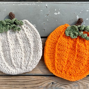 May include: Two crocheted pumpkins, one white and one orange, with green crocheted stems and leaves. The pumpkins are sitting on a wooden surface.