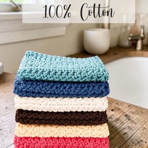 May include: A stack of six square crochet dishcloths made of 100% cotton in teal, blue, white, brown, cream, and pink. The text "Crochet Dishcloths 100% Cotton" is overlaid on the image. Each cloth features a textured, woven stitch pattern.