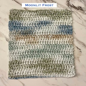 May include: A square crochet washcloth featuring a variegated yarn pattern in shades of sage green, soft blue, white, and tan. The text "MOONLIT FROST" is printed above the textile, which is worked in horizontal rows of double crochet stitches.