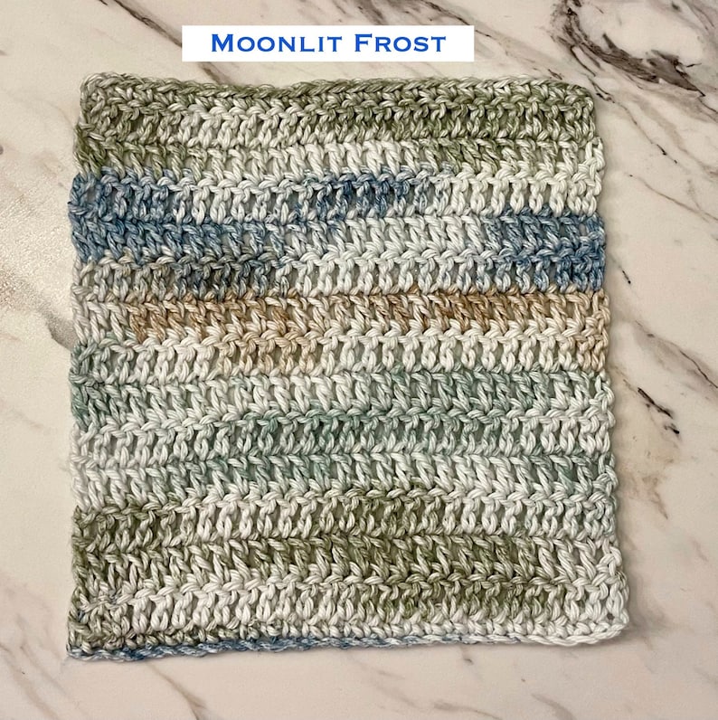 May include: A square crochet washcloth featuring a variegated yarn pattern in shades of sage green, soft blue, white, and tan. The text "MOONLIT FROST" is printed above the textile, which is worked in horizontal rows of double crochet stitches.