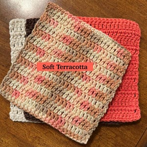 May include: A stack of three square crochet washcloths. The top cloth features a variegated pattern of soft terracotta, cream, and brown. Beneath it lies a solid cream cloth and a solid dark brown cloth, with a solid coral cloth to the side.