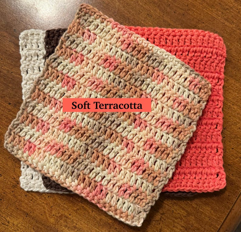 May include: A stack of three square crochet washcloths. The top cloth features a variegated pattern of soft terracotta, cream, and brown. Beneath it lies a solid cream cloth and a solid dark brown cloth, with a solid coral cloth to the side.