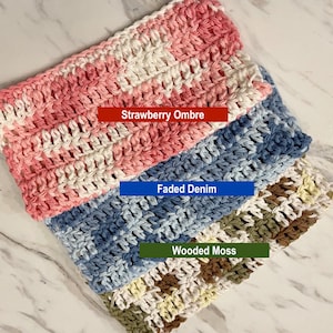 May include: Three rectangular crochet washcloths featuring variegated yarn patterns. The top cloth is pink and white labeled Strawberry Ombre, the middle is blue and white labeled Faded Denim, and the bottom is green, brown, and white labeled Wooded Moss.