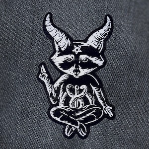 May include: Black and white illustration of a raccoon with devil horns and a pentagram on its forehead, sitting in a meditative pose with its hands in a mudra position. The raccoon is wearing a necklace with a snake design.