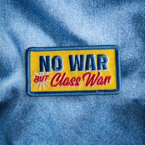 May include: Embroidered patch with the text "NO WAR BUT Class War" in blue and red on a yellow background. The patch has a blue border and a starburst graphic. The patch is on a blue denim background.
